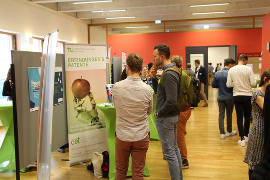 Markt der Möglichkeiten mit Stehtischen und Roll-ups im Gebäudefoyer, Gruppen von Menschen stehen zusammen und unterhalten sich