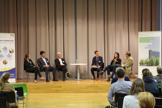 Podiumsdiskussion mit sechs Teilnehmenden (zwei Frauen, vier Männer) auf der Bühne vor Publikum