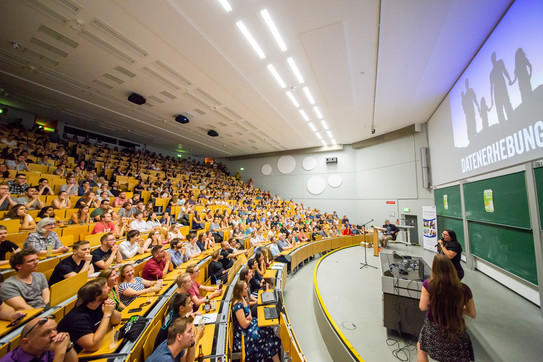 Sehr gut gefüllter Hörsaal mit hölzernen Stuhlreihen, rechts die Bühne mit den Slammerinnen, an der Wand über den Tafeln die projizierte Präsentation