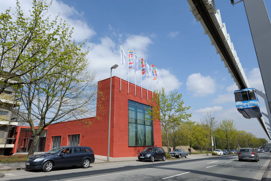 Internationales Begegnungszentrum mit H-Bahn Von Bäumen umgebenes Gebäude des Internationalen Begegnungszentrums und im Hintergrund blauer Himmel mit wenigen Wolken. Rechts im Bild die H-Bahn.