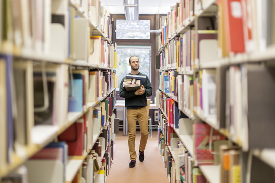 Studierender läuft an den Bücherregalen in der Bibliothek entlang.